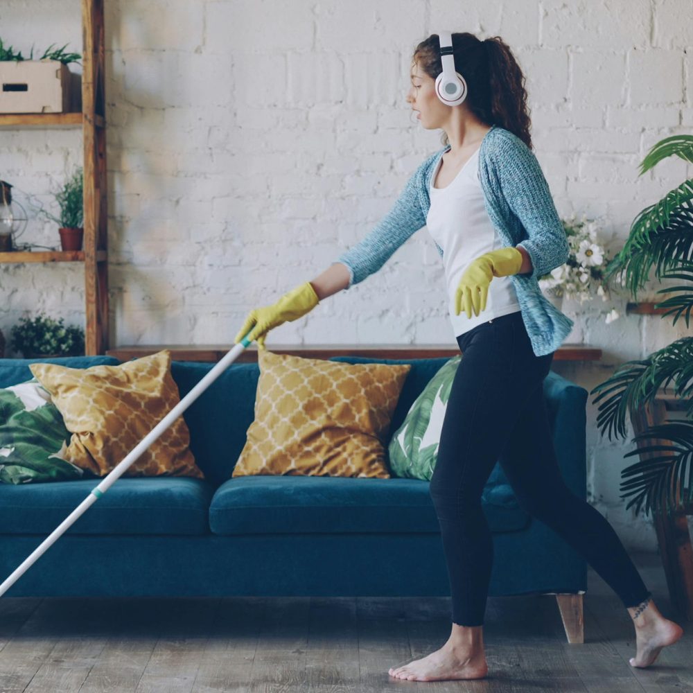 A woman in headphones and yellow rubber gloves mopping the floor in a bright living room
