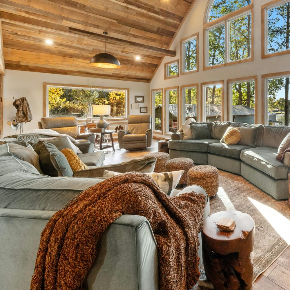 A bright living room with a wood plank ceiling, large windows, sage-green sectional sofas, and a chunky rust-colored knit throw