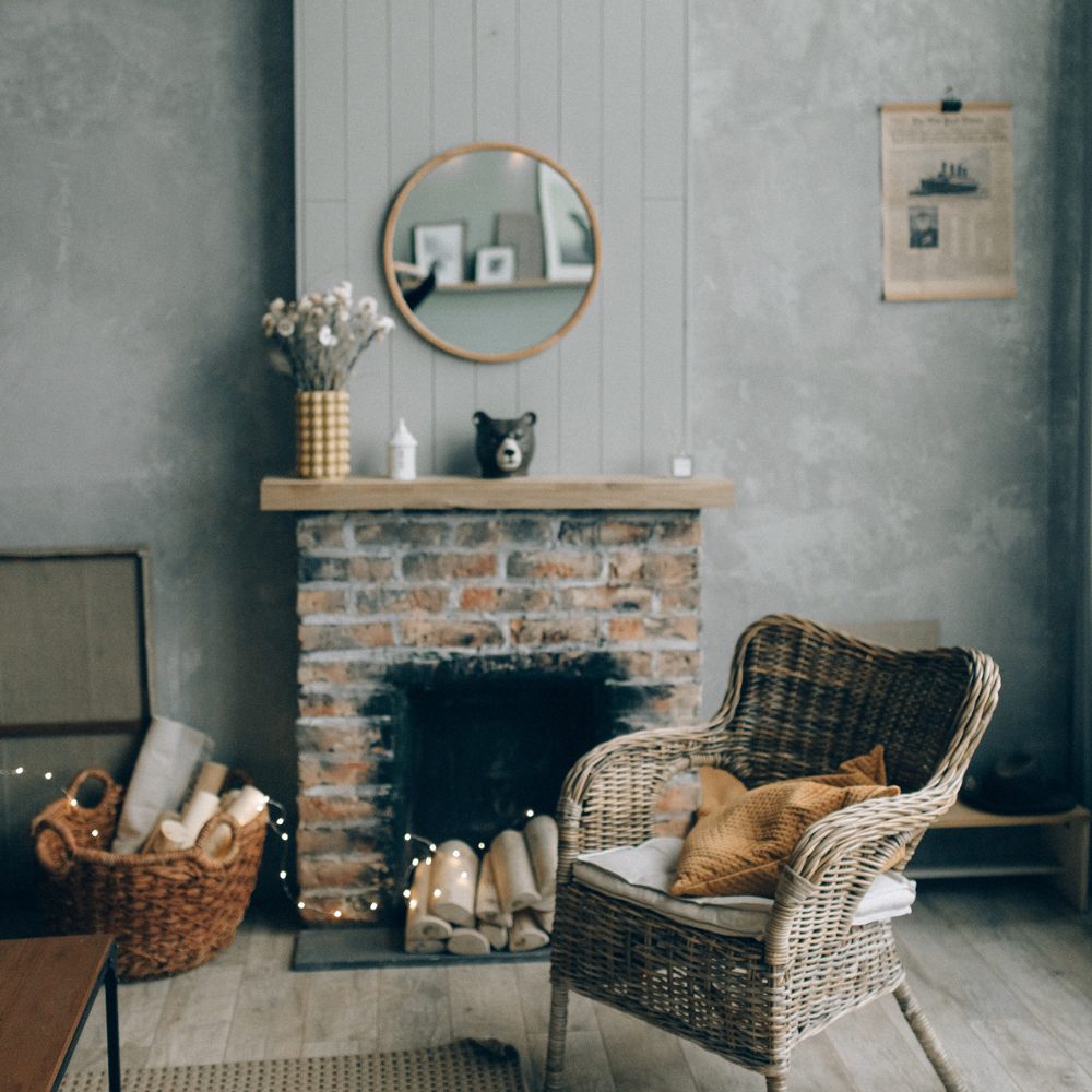 A brick fireplace with a wooden mantel, a round mirror, a wicker chair with a mustard pillow, and a basket of logs