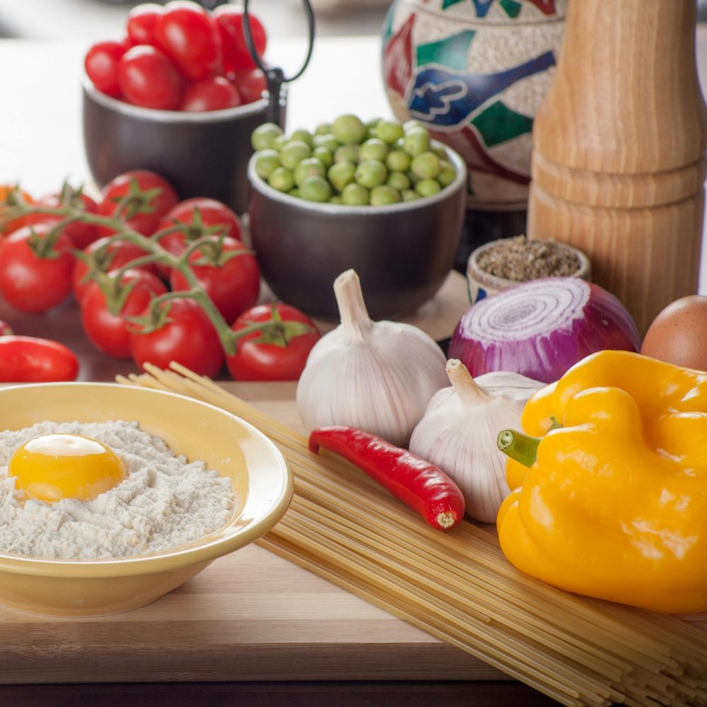 Fresh tomatoes, garlic, an onion, peppers, and spaghetti arranged on a wooden cutting board