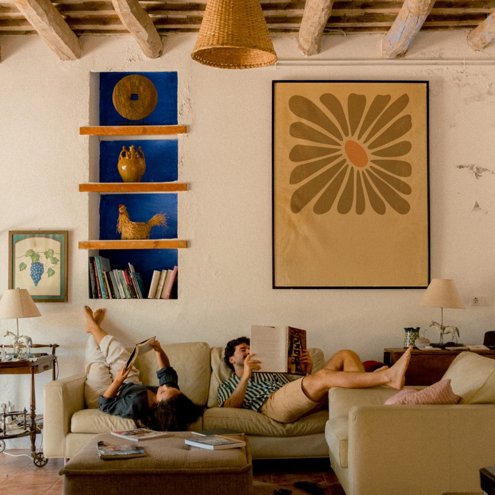 Two people reading on a cream sofa in a warm living room with a large framed floral print and blue shelves